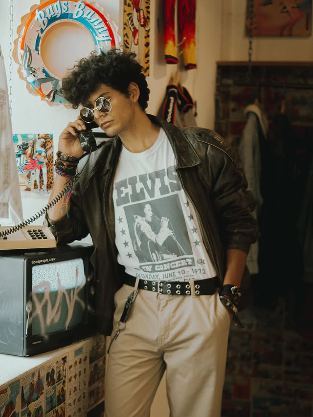 Modelo con lentes redondos hablando por teléfono junto a televisión con grafiti punk